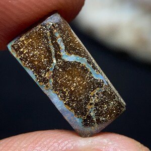 Puede incluir: Una gema rectangular con una superficie marrón moteada y llamativas vetas azules. La piedra tiene un aspecto pulido, con una mezcla de colores y texturas. La gema se sostiene entre dos dedos.