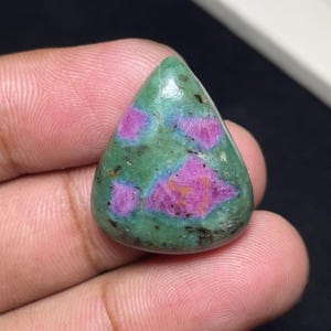 May include: A teardrop-shaped cabochon gemstone featuring a smooth, polished surface with a mottled green base color and distinct, irregular patches of vibrant magenta-pink inclusions throughout the stone, showcasing natural mineral textures.