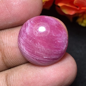 Pode incluir: Um close-up de uma pedra preciosa redonda e polida. A gema é de uma cor rosa vibrante com veios brancos e um acabamento brilhante. A pedra é segurada entre os dedos, com foco na textura e cor da gema.