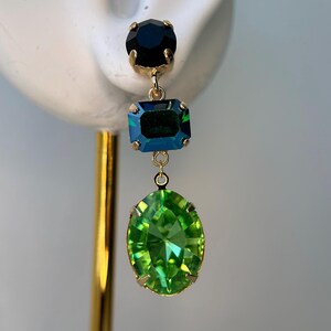 Pode incluir: Um brinco dourado com três pedras preciosas coloridas. A pedra superior é uma pequena gema preta e redonda. A pedra do meio é uma gema retangular azul-esverdeada. A pedra inferior é uma gema grande, oval e verde brilhante.
