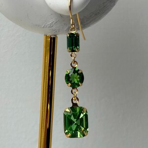 Pode incluir: Um brinco dourado com três pedras verdes. A pedra superior é retangular, a do meio é redonda e a inferior é quadrada. O brinco é suspenso por um gancho dourado.