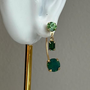 May include: Gold-toned earrings with green gemstones. The earrings feature a round green stone at the bottom, a rectangular green stone in the middle, and a round light green stone at the top. The earrings are displayed on a white ear model.