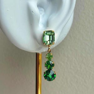 Pode incluir: Brinco dourado com uma pedra retangular verde clara no topo, seguido por três pedras verdes redondas e graduadas. O brinco é exibido em uma forma branca em forma de orelha.