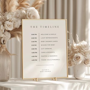 以下が含まれることがあります： 「THE TIMELINE」の文字が入った金色のフレームのサイン。ベビーシャワーのタイムラインが表示され、「ウェルカム＆ミングル」や「ケーキ＆デザート」などのイベントスケジュールが記載されています。白い表面に置かれています。