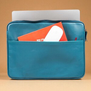 May include: Teal leather laptop sleeve with a front pocket. A silver laptop is inside the sleeve, with an orange booklet and a white mouse in the front pocket. The sleeve has a zipper closure and a small red logo.