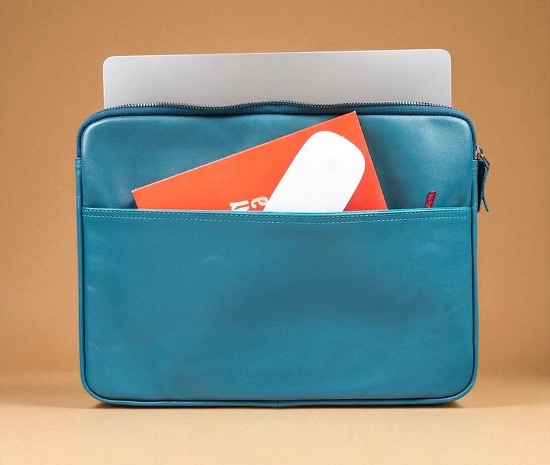 May include: Teal leather laptop sleeve with a front pocket. A silver laptop is inside the sleeve, with an orange booklet and a white mouse in the front pocket. The sleeve has a zipper closure and a small red logo.