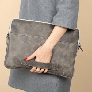 May include: A gray leather laptop sleeve with a white zipper and a handle. The sleeve is rectangular and has a stitched design. The person is wearing a gray dress.