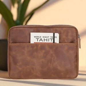 May include: A brown leather tablet case with a front pocket and a zipper closure. A small notebook with the words "WORLD NIGHT TOURS TAHITI" is in the front pocket. The case is rectangular and has a textured appearance.