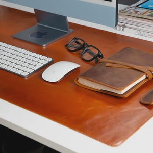 Peut inclure: Un espace de travail avec un tapis de bureau en cuir marron, un clavier, une souris, des lunettes, un journal et un porte-stylo. Un écran d'ordinateur et une pile de magazines sont en arrière-plan. Le bureau est blanc.