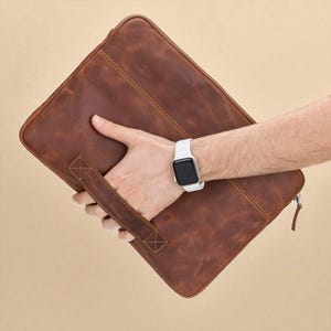 May include: A brown leather laptop sleeve with a handle and zipper closure. The sleeve is held by a person wearing a white smartwatch. The leather has a distressed look with visible stitching.