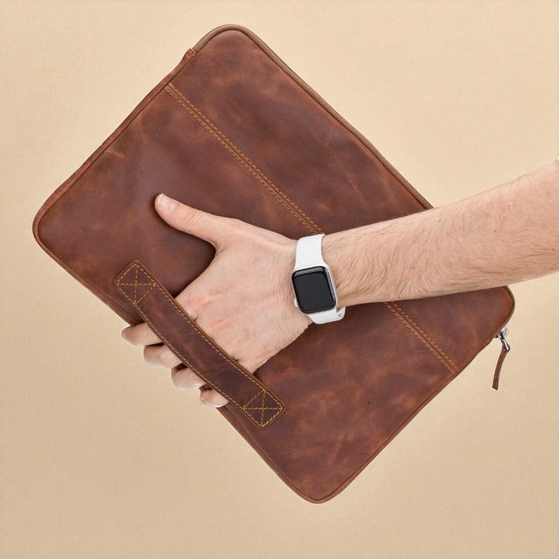 May include: A brown leather laptop sleeve with a handle and zipper closure. The sleeve is held by a person wearing a white smartwatch. The leather has a distressed look with visible stitching.