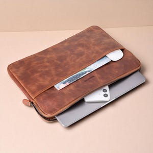 May include: A brown leather laptop sleeve with a zipper closure. The sleeve is open, revealing a laptop, a white mouse, a smartphone, and a colorful card. The leather has a distressed texture.