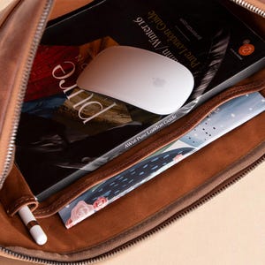 May include: Open brown leather case with a zipper, revealing a white computer mouse, a book titled "Pure London Guide", and a small notebook. A white stylus pen is in a side pocket. The case is filled with various items.