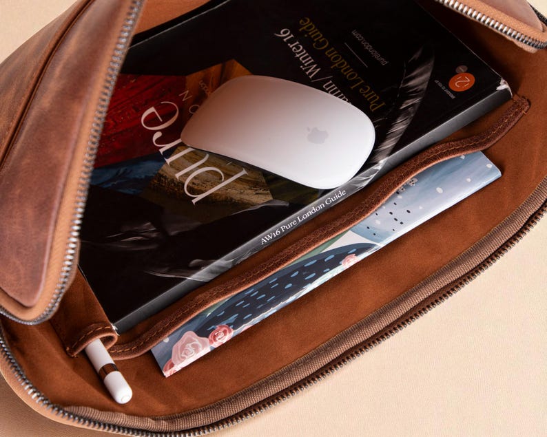 May include: Open brown leather case with a zipper, revealing a white computer mouse, a book titled "Pure London Guide", and a small notebook. A white stylus pen is in a side pocket. The case is filled with various items.