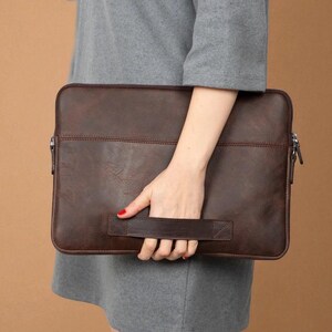 May include: A brown leather laptop sleeve with a handle. The sleeve has a zipper closure and a stitched design. The leather has a textured appearance. The sleeve is held by a person.