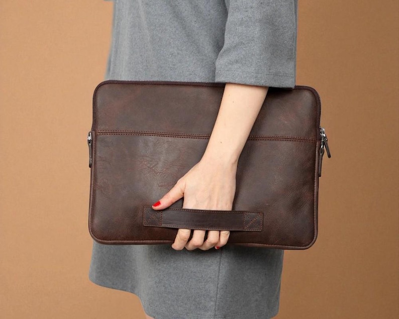 May include: A brown leather laptop sleeve with a handle. The sleeve has a zipper closure and a stitched design. The leather has a textured appearance. The sleeve is held by a person.