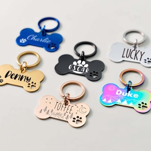 Peut inclure: Plusieurs médailles d'identification pour animaux de compagnie en forme d'os, de différentes couleurs : bleu, doré, noir, blanc et irisé. Chaque médaille porte un nom et des empreintes de pattes. Certaines ont des motifs supplémentaires.