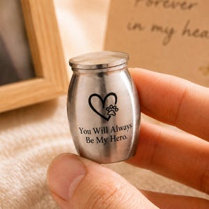 Op de afbeelding: Een kleine, zilverkleurige metalen crematie-urn met een geborstelde afwerking. De voorzijde is voorzien van een zwarte gravure van een hart met een pootafdruk erin. Onder het hart staat de tekst: "You Will Always Be My Hero."