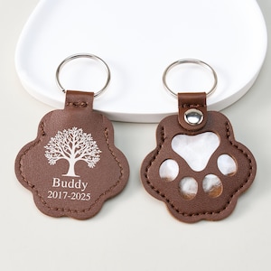 May include: Two brown leather paw-shaped keychains. One keychain features a tree design and the name "Buddy" with the years "2017-2025". The other keychain has clear paw prints. Both have silver key rings.