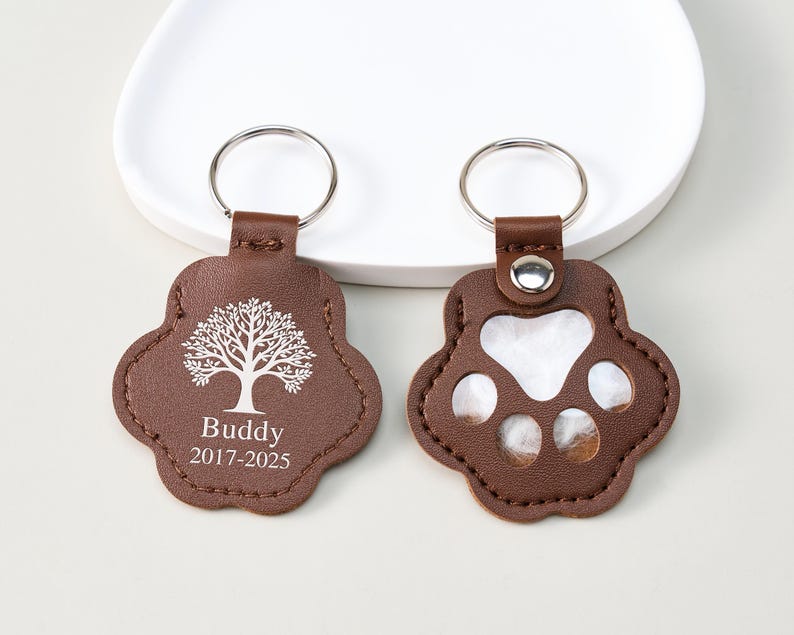 May include: Two brown leather paw-shaped keychains. One keychain features a tree design and the name "Buddy" with the years "2017-2025". The other keychain has clear paw prints. Both have silver key rings.