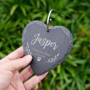 May include: A heart-shaped, dark gray slate memorial stone with the name "Jasper" in elegant script. The text "FOREVER LOVED 2014-2025" and a paw print are also engraved. A silver metal loop is attached for hanging.