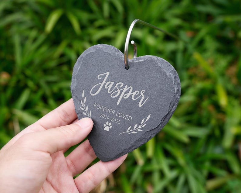 May include: A heart-shaped, dark gray slate memorial stone with the name "Jasper" in elegant script. The text "FOREVER LOVED 2014-2025" and a paw print are also engraved. A silver metal loop is attached for hanging.