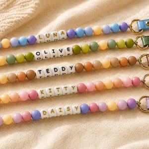 May include: Five personalized beaded lanyards featuring colorful silicone spheres in various pastel and earth tones. Each lanyard includes white letter beads spelling out the names LUNA, OLIVE, TEDDY, KITTY, and DAISY, attached to a leather strap.