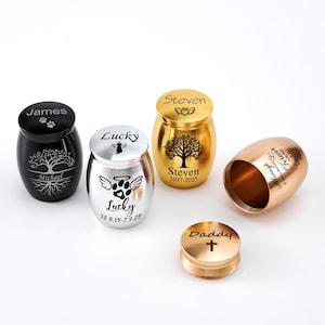 Op de afbeelding: Een verzameling kleine gedenkurnen in zwart, zilver en goud. Elke urn is gegraveerd met namen, data en symbolen, zoals pootafdrukken en bomen. Eén urn is open en toont de binnenkant.