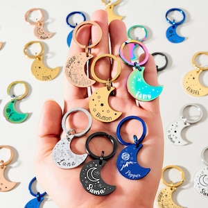 May include: A collection of personalized moon-shaped keychains in various colors, including gold, silver, black, and blue. Each keychain features a name or design, with a keyring attached. The keychains are displayed against a white background.