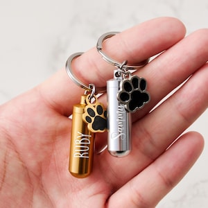 Può includere: Due portachiavi commemorativi per animali domestici in metallo. Uno è dorato con il nome "RUBY" e un ciondolo a forma di zampa nera. L'altro è argentato con il nome "Sawyer" e un ciondolo a forma di zampa nera. Entrambi i portachiavi hanno un portachiavi argentato.