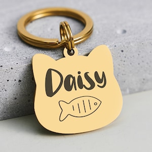 May include: A gold-colored cat-shaped pet tag with the name "Daisy" in black script and a fish illustration. The tag is attached to a gold-colored split ring.
