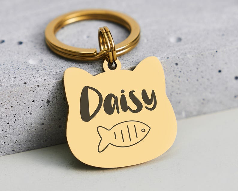 May include: A gold-colored cat-shaped pet tag with the name "Daisy" in black script and a fish illustration. The tag is attached to a gold-colored split ring.