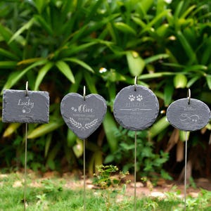 May include: Four dark gray slate pet memorial garden markers with metal stakes. The markers are rectangular, heart-shaped, and oval, with engraved names and paw prints. The text includes "Lucky", "Olive", "Hazel", and "Willow".