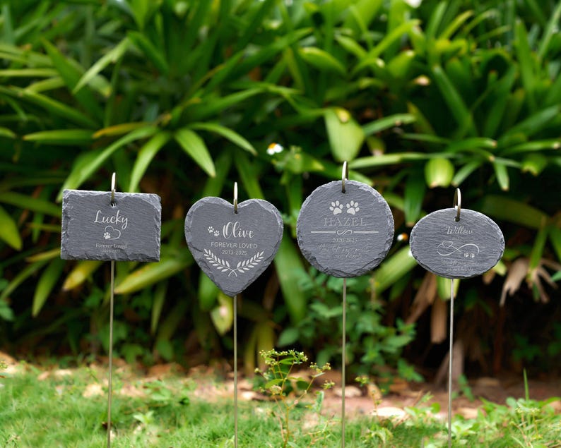 May include: Four dark gray slate pet memorial garden markers with metal stakes. The markers are rectangular, heart-shaped, and oval, with engraved names and paw prints. The text includes "Lucky", "Olive", "Hazel", and "Willow".