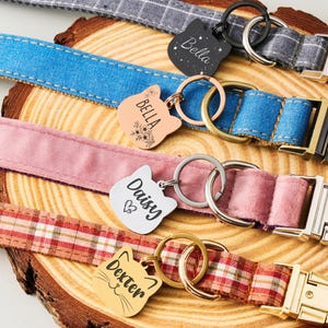 May include: A collection of pet collars in various colors and patterns, including blue denim, pink velvet, and plaid. Each collar features a metal buckle and a cat-shaped tag engraved with a pet's name, such as "Bella" and "Daisy."
