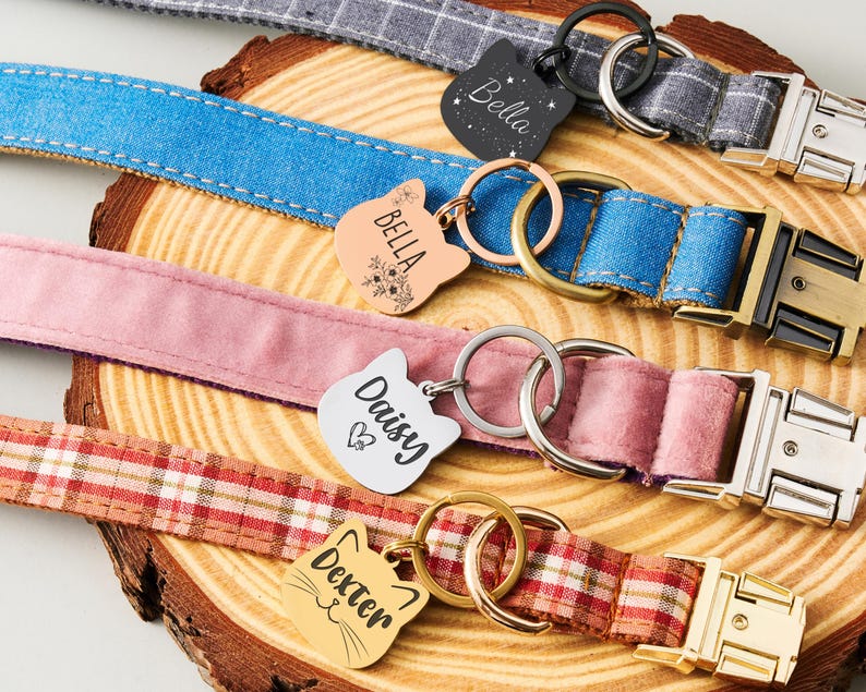 May include: A collection of pet collars in various colors and patterns, including blue denim, pink velvet, and plaid. Each collar features a metal buckle and a cat-shaped tag engraved with a pet's name, such as "Bella" and "Daisy."