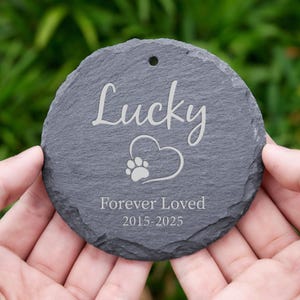 May include: A round, gray slate pet memorial with the name "Lucky" in cursive script. A heart with a paw print is below the name, and the words "Forever Loved 2015-2025" are engraved below. The memorial has a hole at the top.