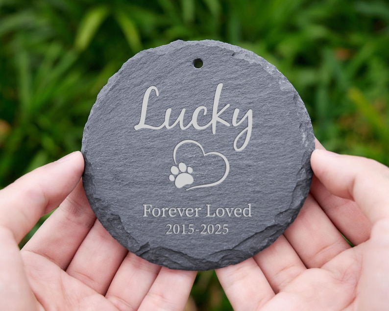 May include: A round, gray slate pet memorial with the name "Lucky" in cursive script. A heart with a paw print is below the name, and the words "Forever Loved 2015-2025" are engraved below. The memorial has a hole at the top.
