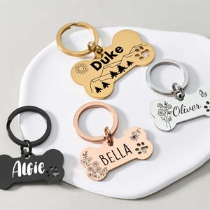 Op de afbeelding: Een verzameling gepersonaliseerde hondenpenningen in de vorm van een bot in goud, roségoud, zilver en zwart. Elke penning heeft een naam en decoratieve elementen zoals bloemen, pootafdrukken en bergontwerpen. De penningen zijn bevestigd aan sleutelringen.