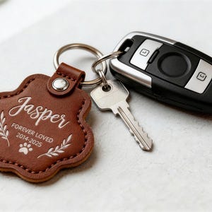 May include: A brown leather paw-shaped keychain with the name "Jasper" and the text "FOREVER LOVED 2014-2025" in white script. The keychain is attached to a silver key ring, a car key fob, and a metal key.