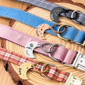 Puede incluir: Primer plano de varios collares para perros con etiquetas con forma de media luna. Los collares son de varios colores y patrones, incluyendo mezclilla, cuadros y rosa liso. Cada etiqueta tiene un nombre grabado, como "Nala" y "Tony".