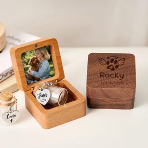 Op de afbeelding: Een houten gedenkdoos voor huisdieren met een foto van een persoon en een hond. De doos is open en toont een kleine glazen pot met een hartvormig label. Een aparte houten doos met een pootafdrukontwerp en de naam "Rocky" is ook zichtbaar.