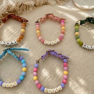 May include: Five pet collars featuring colorful silicone beads and white letter beads spelling out the names TEDDY, KITTY, OLIVE, LUNA, and DAISY. Each collar includes a leather-like strap with a metal buckle and D-ring for secure attachment.