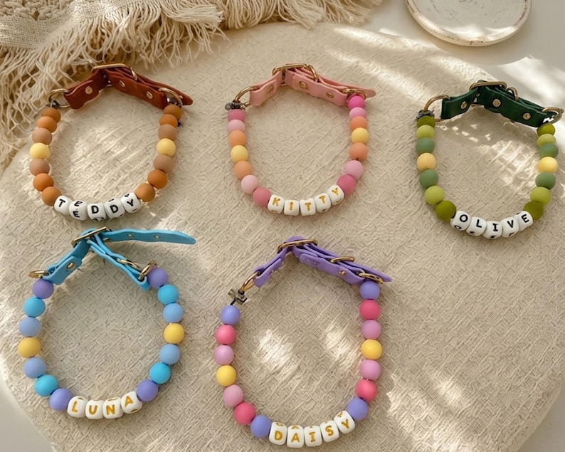 May include: Five pet collars featuring colorful silicone beads and white letter beads spelling out the names TEDDY, KITTY, OLIVE, LUNA, and DAISY. Each collar includes a leather-like strap with a metal buckle and D-ring for secure attachment.