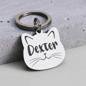 May include: A silver cat-shaped pet tag with the name "Dexter" in black script. The tag has a small loop for attaching to a collar and a silver keyring. The tag is set against a grey, textured background.
