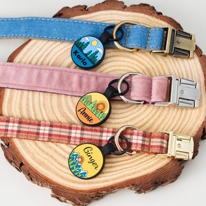 May include: Three dog collars in blue denim, pink velvet, and red plaid patterns, each with a metal buckle and a circular tag. The tags feature landscape designs with names: Karla, Annie, and Ginger.