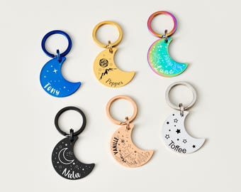 May include: Six crescent moon-shaped keyrings in various colours, each with a different design and name. The keyrings are blue, gold, iridescent, black, rose gold, and white. The names include Tony, Pepper, Luna, Nala, Vanille, and Toffee.