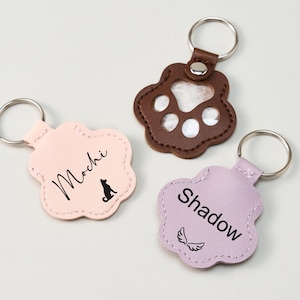 May include: Three paw-shaped keychains in pink, brown, and lavender. The pink keychain has the name "Mochi" and a dog silhouette. The brown keychain has a paw print design. The lavender keychain has the name "Shadow" and angel wings.