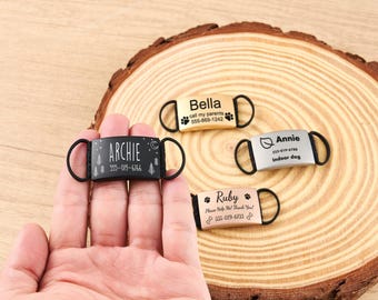 May include: A hand holding a black dog tag with the name "Archie", a phone number, and a forest design. Other tags include "Bella", "Annie", and "Ruby", each with a name and contact information. The tags have black loops.