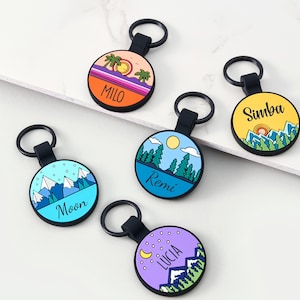 May include: A selection of personalised keyrings with black straps and key rings. Each keyring has a colourful circular design with a name and a landscape illustration, including mountains, trees, and a sunset. The names on the keyrings are Milo, Simba, Remi, Moon, and Lucia.